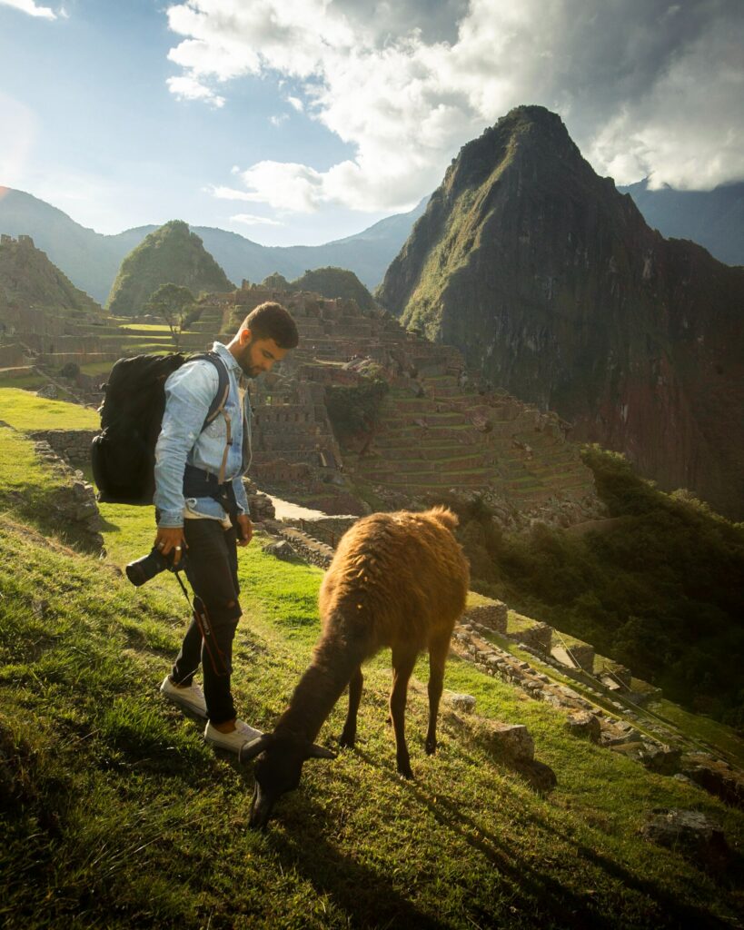 traveling man with a camera standing with a lama with green mountains and ancient ruins behind him.