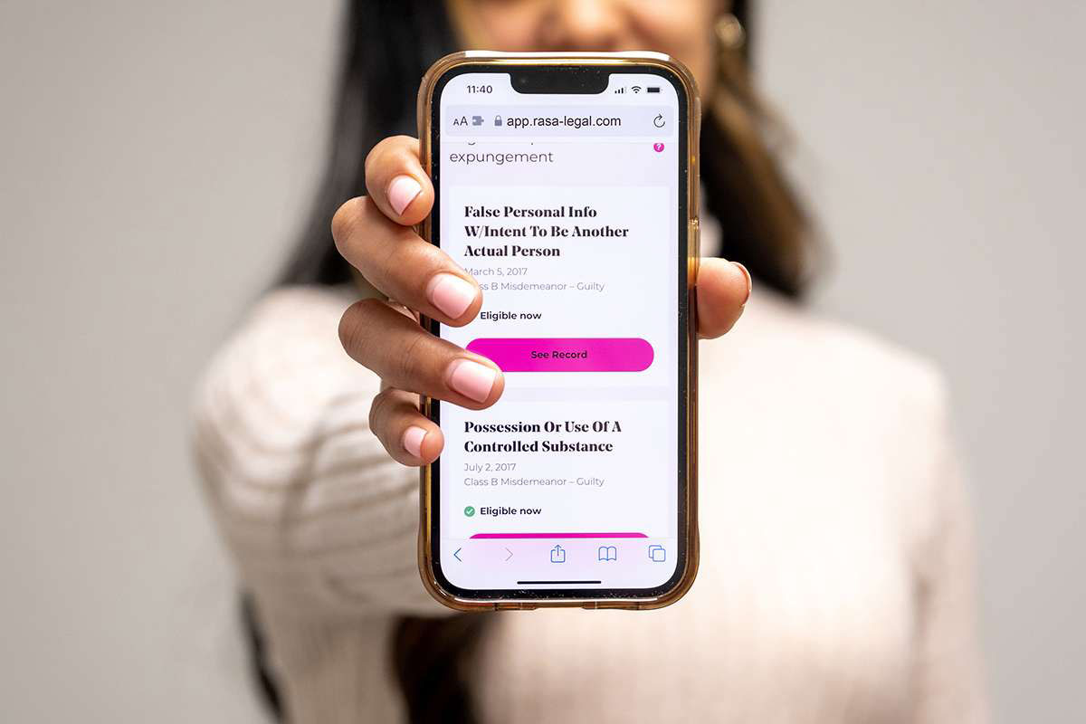 29112358 A person holds a smartphone displaying an expungement eligibility app. The screen shows two criminal records: 'False Personal Info W/Intent To Be Another Actual Person' dated March 5, 2017, and 'Possession Or Use Of A Controlled Substance' dated July 2, 2017. Both are listed as Class B Misdemeanors with 'Guilty' verdicts and are marked 'Eligible now' for expungement. The app interface includes a pink 'See Record' button. The phone's address bar shows 'app.rasa-legal.com'. Only the person's hands and part of their white clothing are visible in the image.