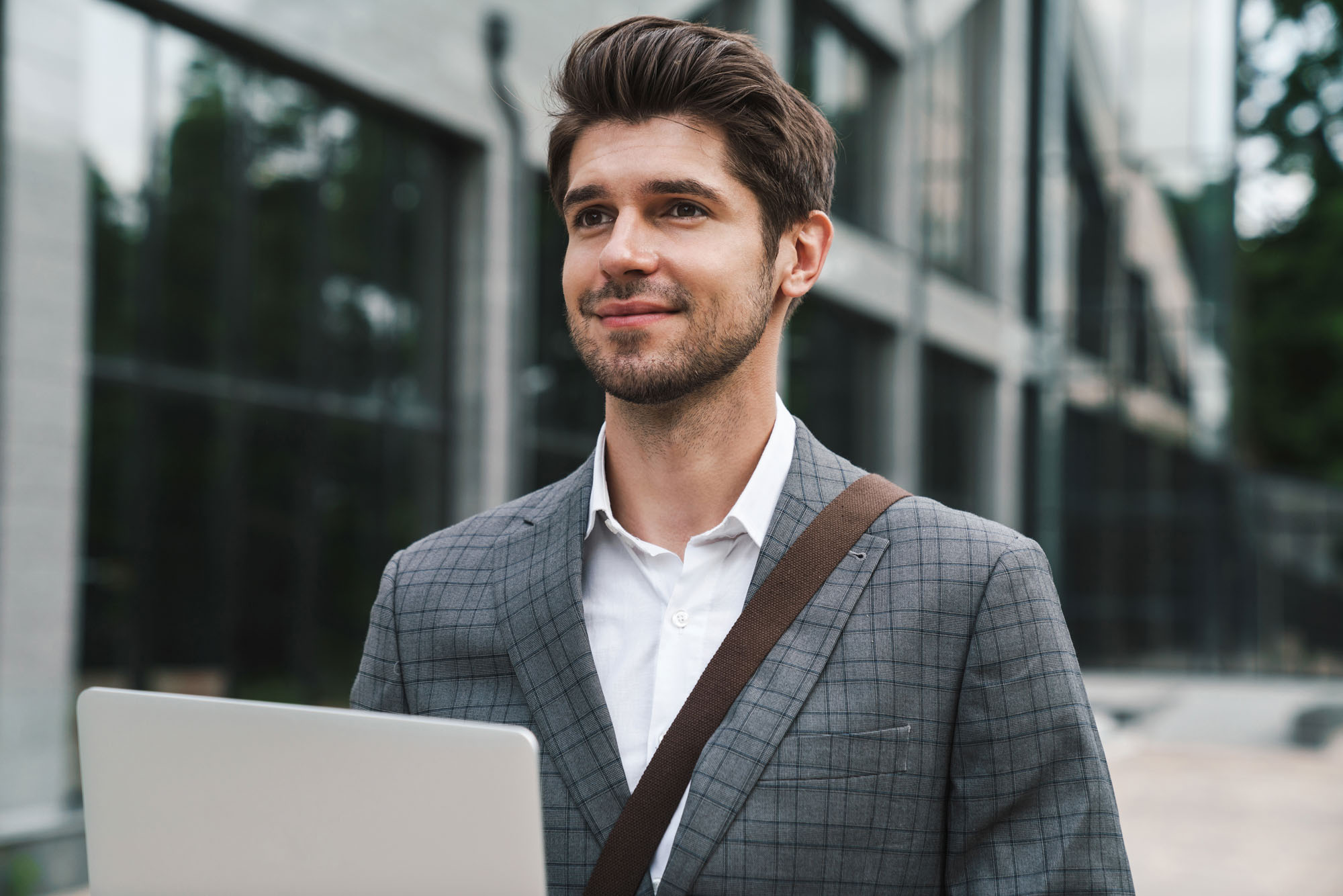 Image of pleased optimistic handsome business man outdoors using laptop computer near business center Image of pleased optimistic handsome business man outdoors using laptop computer near business center